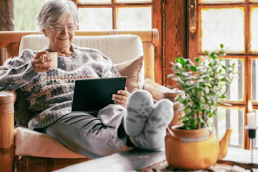 Older lady wearing glasses, a heavy sweater, slacks, and heavy socks, sitting comfortably in a chair, sipping coffee or tea, and ready from an electronic tablet. 